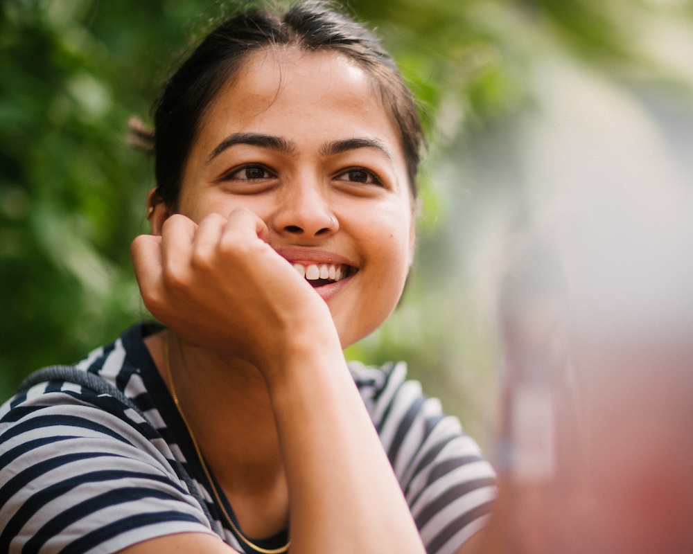 Candid close-up portrait of a woman smiling with her chin resting on her hand, shot in natural light with shallow depth of field and lush green bokeh at La Canopée (Eaux-Vives) in Geneva.