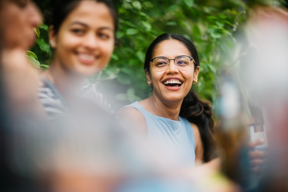 Candid and cinematic portrait of a woman with glasses laughing joyfully at an outdoor gathering, framed by soft foreground bokeh and lush green foliage at Plage des Eaux-Vives in Geneva.