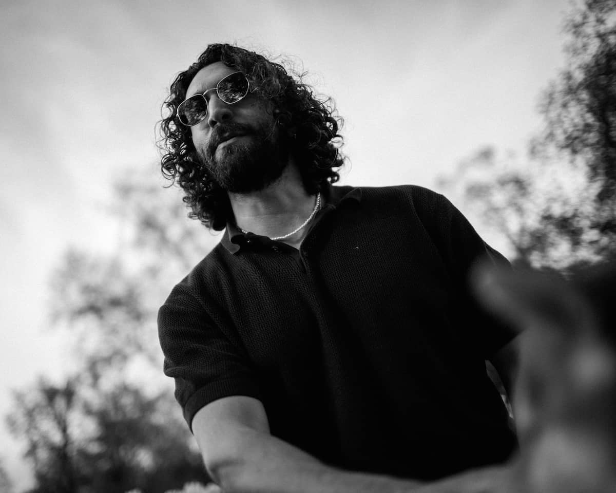 Low angle black and white cinematic portrait of a bearded man with curly hair and sunglasses against a bright sky at Perle du Lac in Geneva