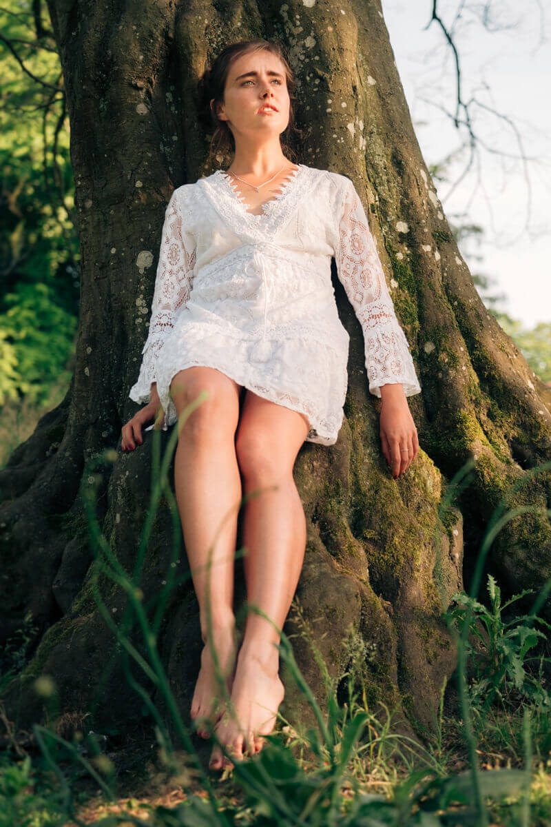 Cinematic outdoor portrait of a young woman in a white lace dress leaning against a large mossy tree trunk at Parc La Grange in Geneva during golden hour.