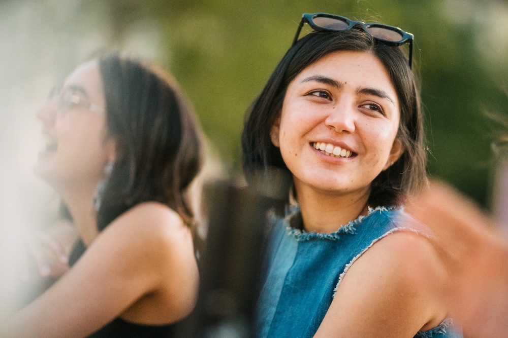 A spontaneous cinematic portrait of a woman smiling outdoors during golden hour at the Geneva Street Food Festival in Plainpalais, framed by soft foreground flares and a laughing friend