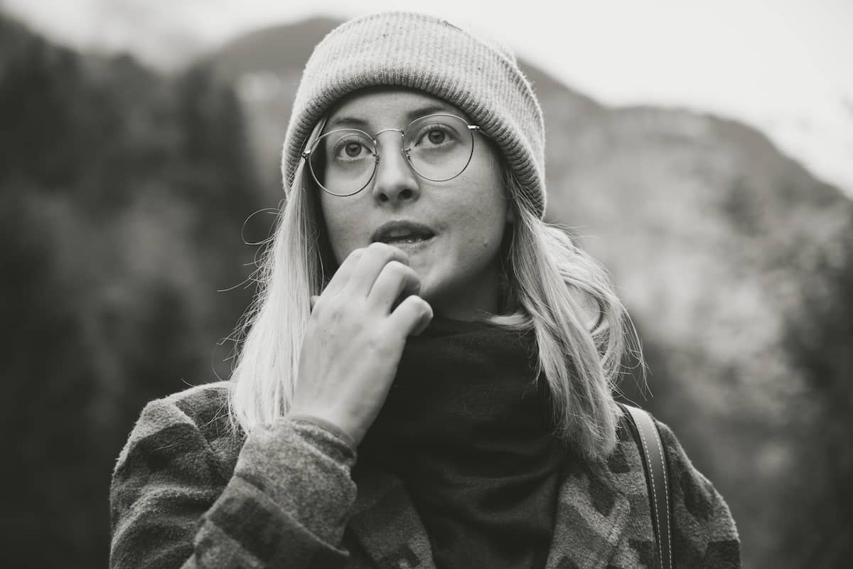 Close-up black and white cinematic portrait of a young woman with round glasses and a beanie, looking contemplatively upwards in a natural Swiss mountain environment in the region of Lauterbrunnen.
