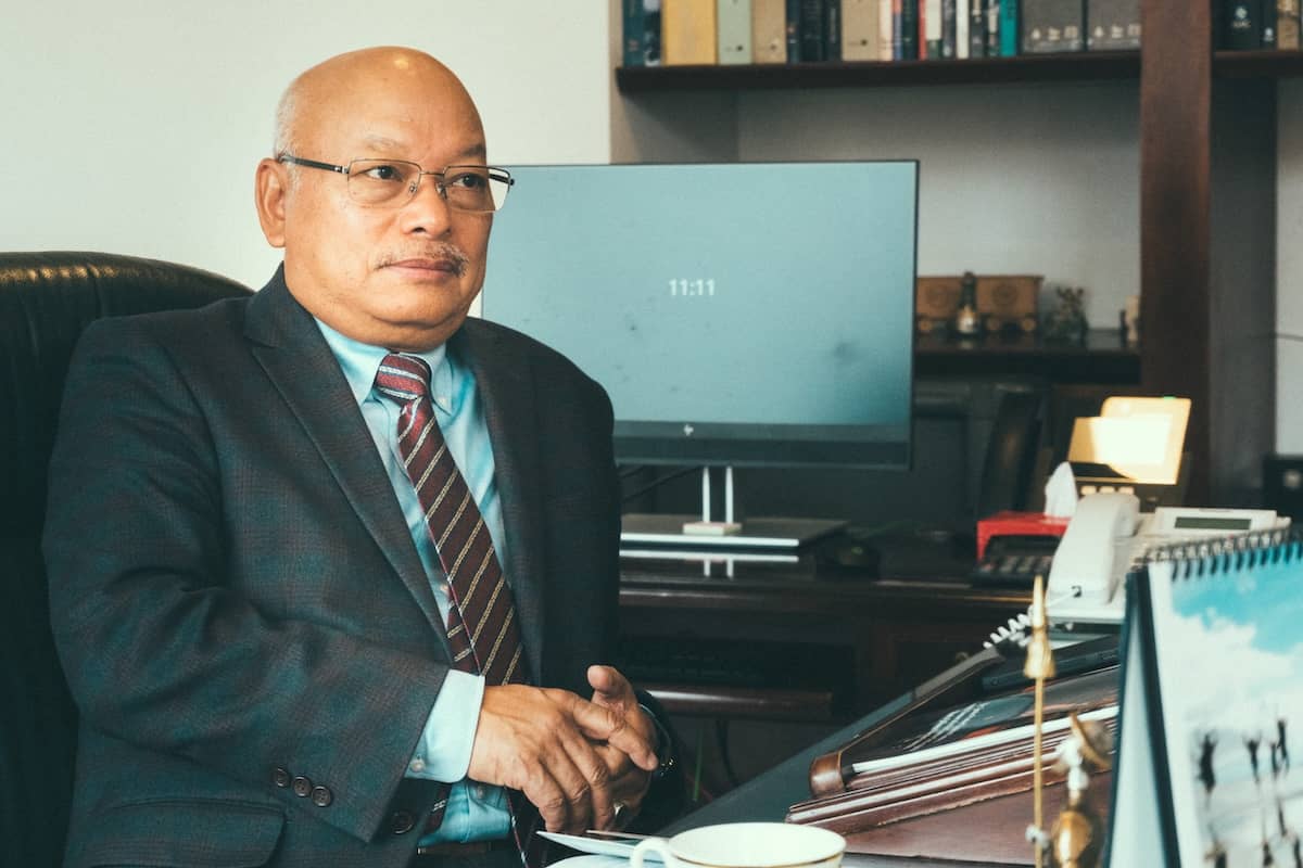 Candid documentary portrait of an ambassador in a suit sitting inside his office at the Embassy of India in Bogota, looking thoughtfully out a window with official documents on a table nearby.