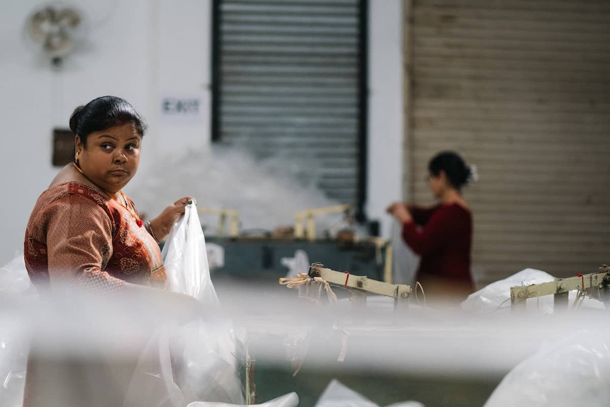 Candid documentary portrait of a woman in a patterned terracotta dress working in an industrial setting, looking thoughtfully to the side while holding white plastic material.