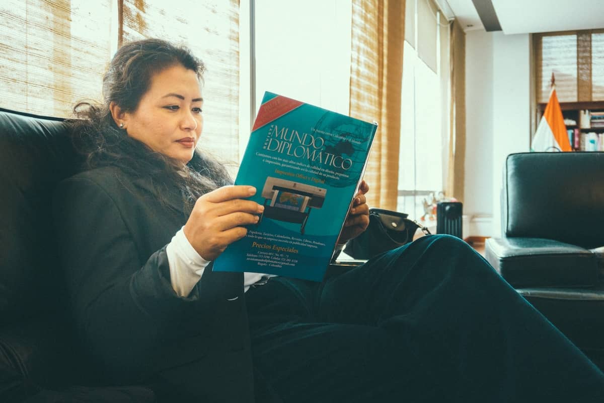 Candid editorial portrait of a woman at the Embassy of India in Bogota in a dark blazer sitting on a leather sofa and reading a magazine titled Mundo Diplomatico in a bright room with window light.