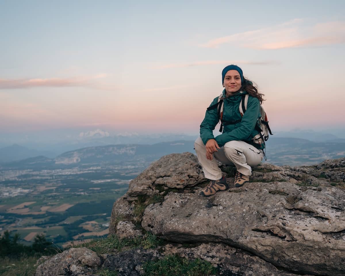 Wide cinematic portrait of a woman in hiking gear crouching on a rocky mountain peak at sunset with the Jura mountains and Geneva valley in the background.