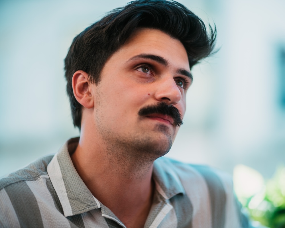 Cinematic candid portrait of a man looking upward with a thoughtful expression, shot wide-open with an organic focus roll-off wrapping the highlights on his striped shirt and a creamy, beautifully dissolved background.